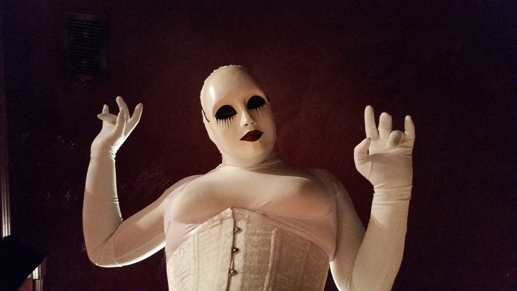 A photo of evil dolly clad all in white girdles, a white corset, and a white creepy doll mask. She's looking down at the camera, hands lifted in delight. 