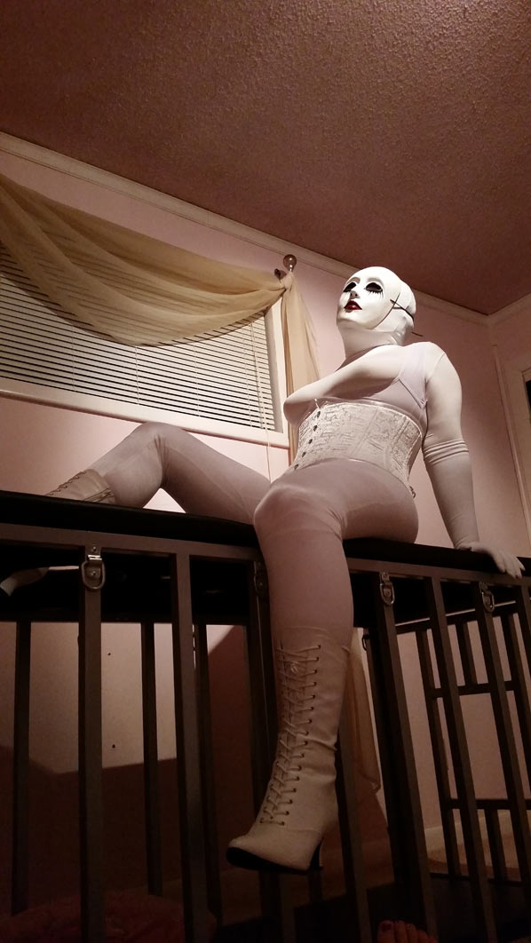A photo of evil dolly clad all in white girdles, a white corset, white boots, and a white creepy doll mask. She's sitting on top of a cage.
