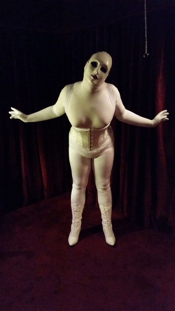 A photo of evil dolly clad all in white girdles, a white corset, and a white creepy doll mask. She's posing for the camera in a room with wine colored curtains and carpet.