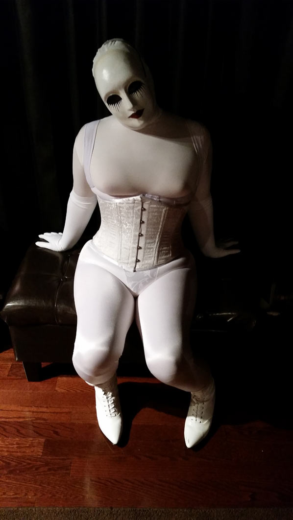 A photo of evil dolly clad all in white girdles, a white corset, and a white creepy doll mask. She's sitting on a black stool. POV from above.