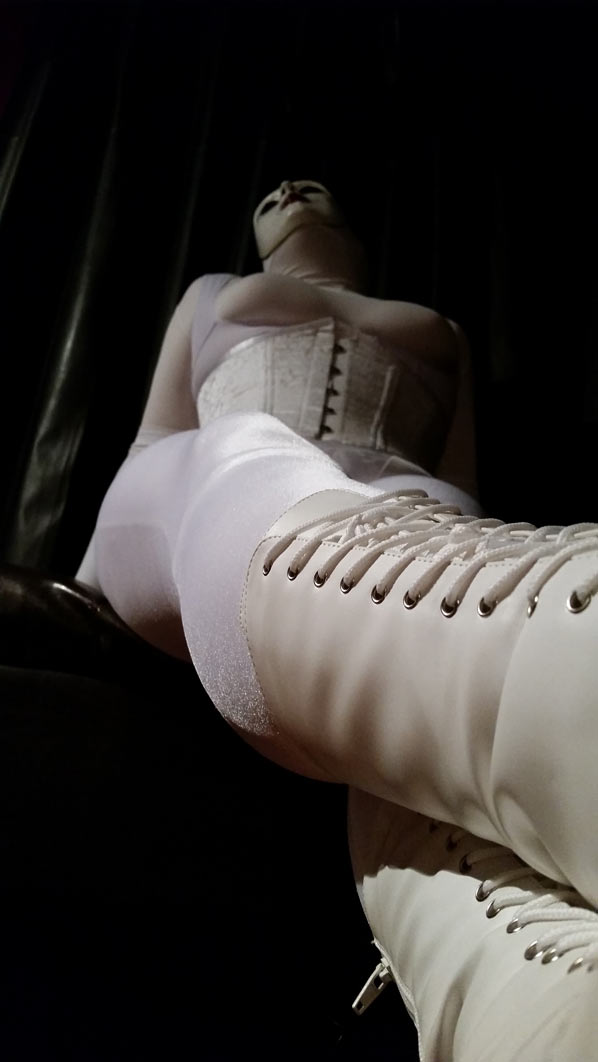 A photo of evil dolly clad all in white girdles, a white corset, and a white creepy doll mask. She's sitting and looking up. POV from her boots.