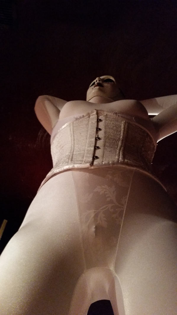 A photo of evil dolly clad all in white girdles, a white corset, and a white creepy doll mask. She's stretching and looking up. POV from below.