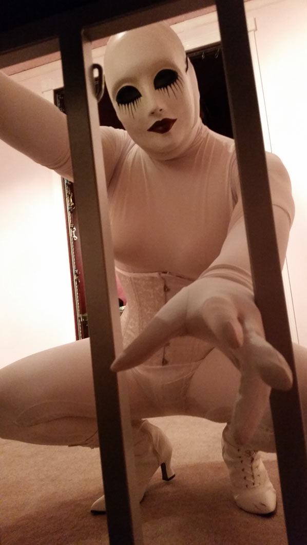A photo of evil dolly clad all in white girdles, a white corset, and a white creepy doll mask. She's reaching into a cage through the bars as if to grab the viewer.